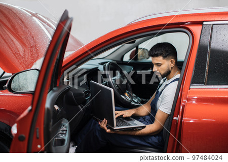 Bearded male mechanic sitting inside car using laptop, recording automobile engine checks Bearded male mechanic sitting inside car using laptop, recording automobile engine checks 97484024