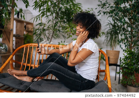 Side view of cheerful young African American woman listening music from mobile phone, choosing next song sitting on couch at home in room with modern green biophilic interior design. 97484492