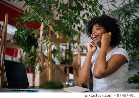 Low-angle view of joyful excited African American female student listening music in headphones sitting at table with laptop and smiling looking away, in light home office room with biophilic interior. 97484493