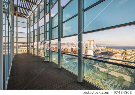 Long spacious terrace in high-rise building with fence and roof made of metal beams of strong impact-resistant glass protected from direct sunlight. Floor of terrace is covered with wooden parquet. 97484508