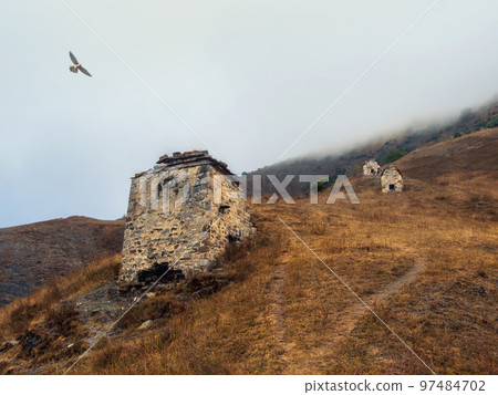 A hiking trail uphill through ancient family crypts, a misty mystical landscape. Old Erzi tower complex in the Jeyrah gorge, located on the extremity of the mountain range in Ingushetia, Russia. A hiking trail uphill through ancient family crypts, a misty mystical landscape. Old Erzi tower complex in the Jeyrah gorge, located on the extremity of the mountain range in Ingushetia, Russia. 97484702