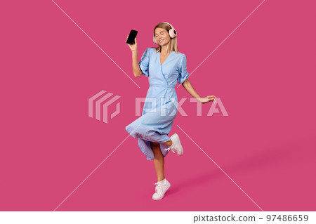 Cheerful mature caucasian female blonde in dress and wireless headphones shows smartphone with empty screen 97486659