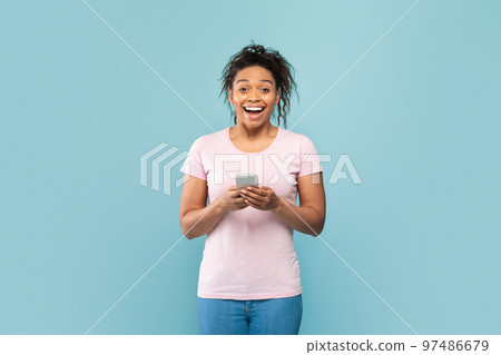 Surprised african american lady holding smartphone and looking at camera in excitement, blue studio background Surprised african american lady holding smartphone and looking at camera in excitement, blue studio background 97486679