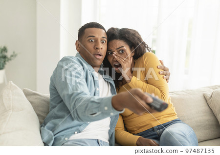Scared shocked young african american woman and man with remote control switch channel on tv, watch horror film 97487135