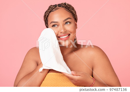 Attractive african american body positive woman drying face with soft towel, caring for skin over pink background 97487136