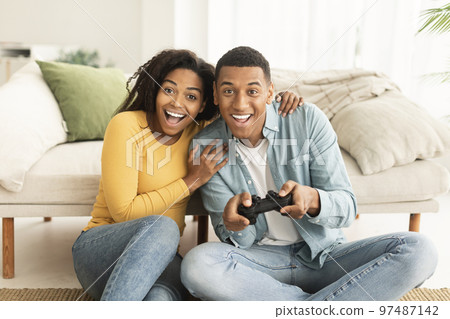 Happy excited young african american wife and husband sit on floor with joystick, play in online game Happy excited young african american wife and husband sit on floor with joystick, play in online game 97487142