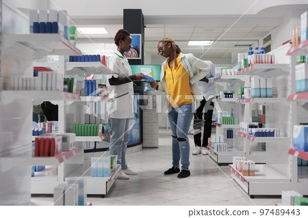 Pharmacy consultant helping buyer choosing back pain medication, side view wide shot. African american woman client with backache standing in drugstore aisle, shopping, talking with pharmacist 97489443