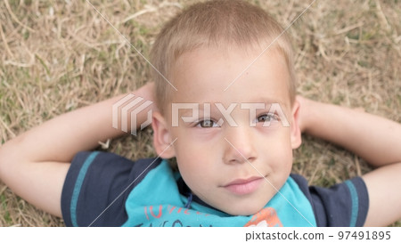 Little Happy Serious and Thoughtful Child Boy Laying on Yellow Lawn Dry Grass Hay in Park. Summer Time, Nature, Dreams, Lifestyle Country life farm village. Smilling Face Close up Looking at Camera 97491895