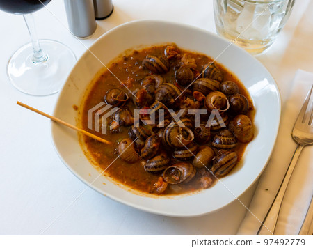 Stewed snails in red sauce served on a white ceramic plate close to the cutlery and bread in the restaurant Stewed snails in red sauce served on a white ceramic plate close to the cutlery and bread in the restaurant 97492779