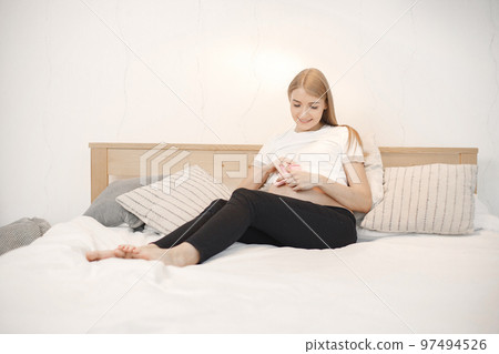 Portrait of pregnant woman holding a little baby rose socks. Pregnant woman lie on a bed in living room. Blonde woman wearing white t-shirt and black trousers. Portrait of pregnant woman holding a little baby rose socks. Pregnant woman lie on a bed in living room. Blonde woman wearing white t-shirt and black trousers. 97494526