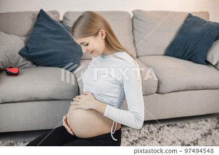Portrait of pregnant woman touching her belly. Pregnant woman sitting on a sofa in living room. Blonde woman wearing grey sweater. Portrait of pregnant woman touching her belly. Pregnant woman sitting on a sofa in living room. Blonde woman wearing grey sweater. 97494548