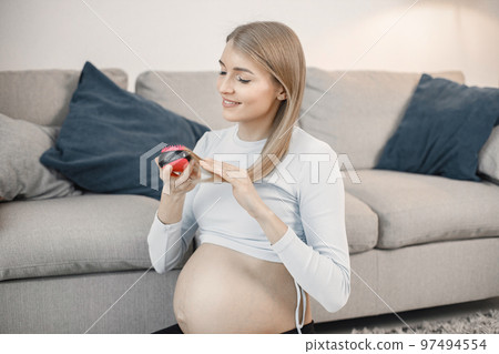 Portrait of pregnant woman brushing her hair. Pregnant woman sitting on a sofa in living room. Blonde woman wearing grey sweater. Portrait of pregnant woman brushing her hair. Pregnant woman sitting on a sofa in living room. Blonde woman wearing grey sweater. 97494554