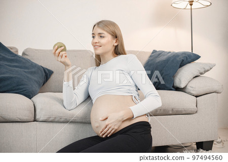 Portrait of pregnant woman holding an apple. Pregnant woman sitting on a sofa in living room. Blonde woman wearing grey sweater. Portrait of pregnant woman holding an apple. Pregnant woman sitting on a sofa in living room. Blonde woman wearing grey sweater. 97494560