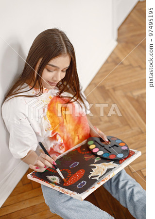 Young female artist posing holding brushes and painting in hands. Woman wearing white t-shirt and blue jeans. Brunette girl sitting in a chair in room with white wall. Young female artist posing holding brushes and painting in hands. Woman wearing white t-shirt and blue jeans. Brunette girl sitting in a chair in room with white wall. 97495195