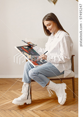Satisfied female artist posing holding new picture and paint palette in hands. Woman wearing jeans and white shirt. Brunette girl sitting on a chair in room with white wall. Satisfied female artist posing holding new picture and paint palette in hands. Woman wearing jeans and white shirt. Brunette girl sitting on a chair in room with white wall. 97495247