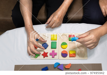 Caregiver and senior woman playing wooden shape puzzles game for dementia prevention 97495942