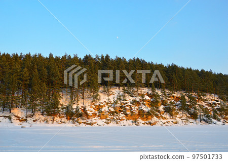 Winter landscape with the moon in the sky during the day. The river is covered with snow. Pine trees grow on red stones. Nature of Eastern Siberia. 97501733