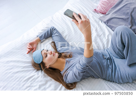 Beautiful woman in blue pajamas looking into phone. Morning starts with phone. 97503850