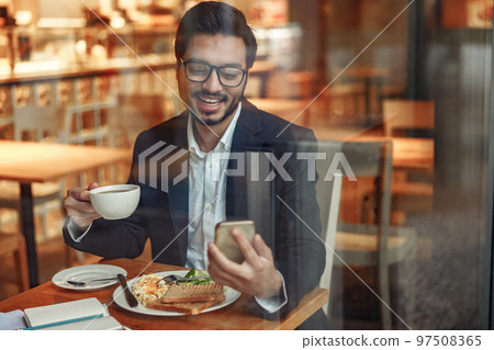 Smiling Indian businessman wearing suit is using phone during lunch time in cafe. Blurred background Smiling Indian businessman wearing suit is using phone during lunch time in cafe. Blurred background 97508365