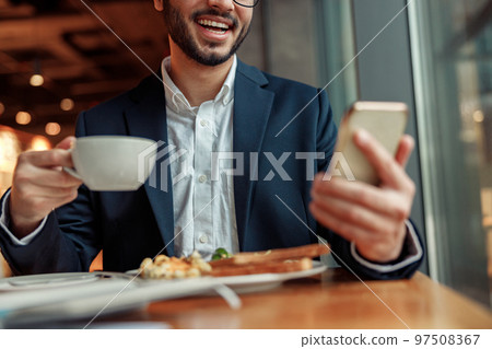 Smiling Indian businessman wearing suit is using phone during lunch time in cafe. Blurred background Smiling Indian businessman wearing suit is using phone during lunch time in cafe. Blurred background 97508367
