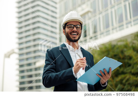 Indian construction architect in suit and hardhat making notes against modern building 97508498