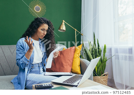 Frustrated and sad woman at home paying bills and paying mortgage, hispanic woman has no money for paperwork using laptop and calculator to calculate household budget sitting on sofa. 97512485