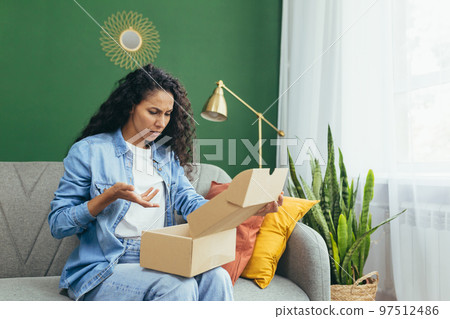 Dissatisfied woman buyer received wrong parcel and wrong product, cheated latina woman sitting on sofa at home in living room. Dissatisfied woman buyer received wrong parcel and wrong product, cheated latina woman sitting on sofa at home in living room. 97512486