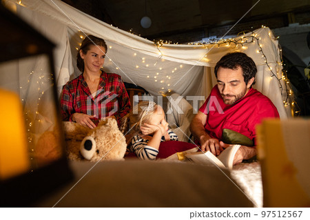 Happy family, mother, father and daughter lying inside hut, tent with toys, pillows and Christmas lights. Cozy evening 97512567