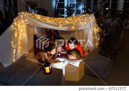 Beautiful family, mother, father and daughter lying inside self-made hut, tent in room in the evening and reading book with flashlight 97512577