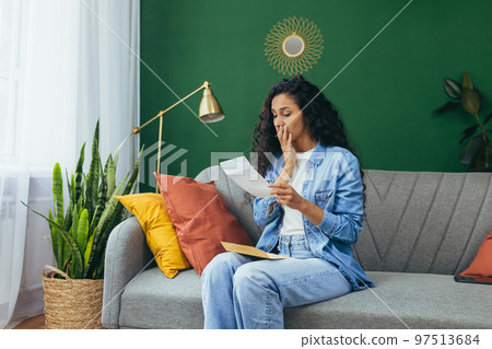 Sad woman at home reading bad news from letter received by mail, Hispanic woman sitting on sofa in living room. 97513684
