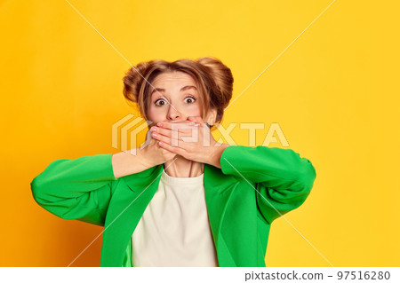 Portrait of young girl in bright green jacket posing, covering mouth with hands over yellow studio background. Secrets 97516280