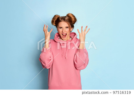 Portrait of young emotional girl in pink hoodie posing with irritated, angry face over blue studio background. Bad vibes 97516319