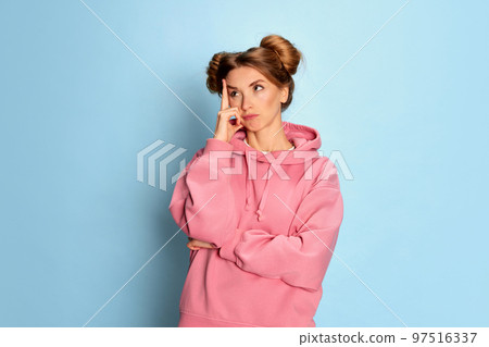 Portrait of young girl in pink hoodie posing with thoughtful face over blue studio background. Creativity and brainstorming Portrait of young girl in pink hoodie posing with thoughtful face over blue studio background. Creativity and brainstorming 97516337