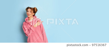 Portrait of young positive girl in pink hoodie posing, pointing with excitement over blue studio background. Good news. Flyer 97516367