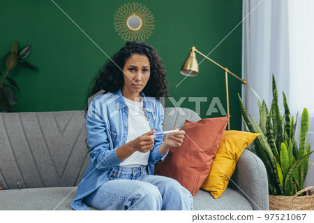 Portrait of a woman at home, unhappy hispanic woman with curly hair looking at the camera and holding an unwanted pregnancy test. Portrait of a woman at home, unhappy hispanic woman with curly hair looking at the camera and holding an unwanted pregnancy test. 97521067