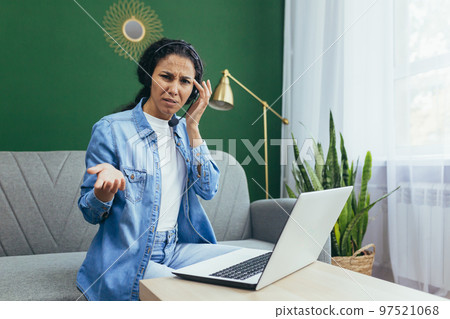 Portrait of frustrated and upset woman at home, Hispanic woman working remotely looking at laptop at work and video call headset sitting on sofa in living room. Portrait of frustrated and upset woman at home, Hispanic woman working remotely looking at laptop at work and video call headset sitting on sofa in living room. 97521068