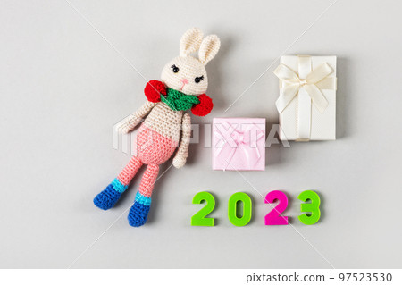 Knitted children's toy bunny or rabbit with pink and beige gift boxes and numbers 2023 on a gray background. The symbol of 2023. 97523530