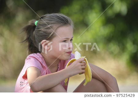 Young pretty child girl eating tasty ripe banana snacking outdoors on summer day Young pretty child girl eating tasty ripe banana snacking outdoors on summer day 97525429