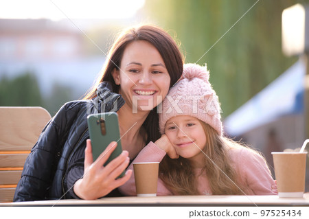 Young mother and her child daughter taking picture with phone selfie camera sitting at street cafe with hot drinks on sunny fall day. Social media presence in everyday life concept 97525434