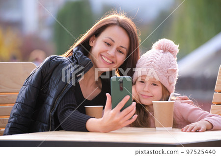 Young mother and her child daughter taking picture with phone selfie camera sitting at street cafe with hot drinks on sunny fall day. Social media presence in everyday life concept Young mother and her child daughter taking picture with phone selfie camera sitting at street cafe with hot drinks on sunny fall day. Social media presence in everyday life concept 97525440