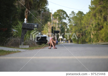 Young moody child girl squatting in the middle of the road covering face with hands on sunny day Young moody child girl squatting in the middle of the road covering face with hands on sunny day 97525448