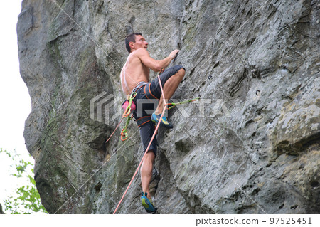 Young man climbing steep wall of rocky mountain. Male climber overcomes challenging route. Engaging in extreme sport concept. Young man climbing steep wall of rocky mountain. Male climber overcomes challenging route. Engaging in extreme sport concept. 97525451