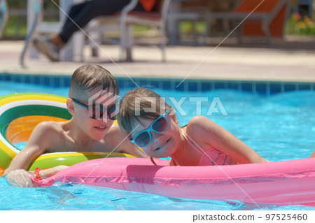 Young joyful children boy and girl having fun swimming on inflatable air mattress in swimming pool with blue water on warm summer day on tropical vacations. Summertime activities concept Young joyful children boy and girl having fun swimming on inflatable air mattress in swimming pool with blue water on warm summer day on tropical vacations. Summertime activities concept 97525460