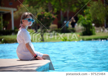 Young joyful child girl in white dress resting on swimming pool side with clear blue water on sunny summer day. Tropical vacations concept Young joyful child girl in white dress resting on swimming pool side with clear blue water on sunny summer day. Tropical vacations concept 97525464
