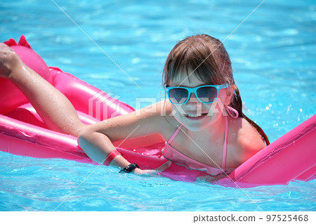 Young joyful child girl having fun swimming on inflatable air mattress in swimming pool with blue water on warm summer day on tropical vacations. Summertime activities concept Young joyful child girl having fun swimming on inflatable air mattress in swimming pool with blue water on warm summer day on tropical vacations. Summertime activities concept 97525468
