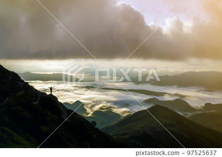 Wide mountain panorama. Small silhouette of tourist with backpack on rocky mountain slope with raised hands over valley covered with white puffy clouds. Beauty of nature, tourism and traveling concept Wide mountain panorama. Small silhouette of tourist with backpack on rocky mountain slope with raised hands over valley covered with white puffy clouds. Beauty of nature, tourism and traveling concept 97525637