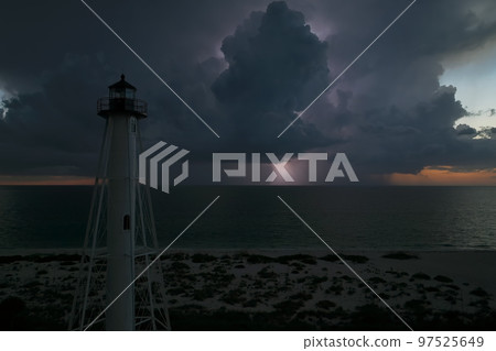 White tall lighthouse on sea shore with blinking light at stormy night for commercial vessels navigation. Thunderstorm with lightnings over ocean water posing danger for ships White tall lighthouse on sea shore with blinking light at stormy night for commercial vessels navigation. Thunderstorm with lightnings over ocean water posing danger for ships 97525649