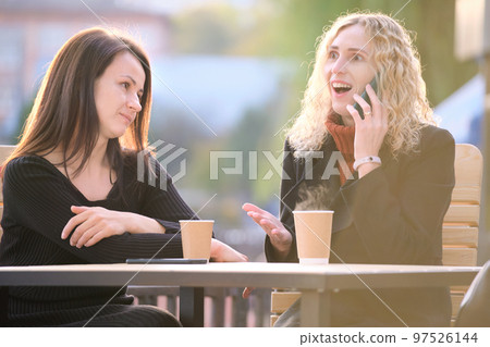 Unhappy young woman waiting angrily while her friend is talking happily on sellphone with someone else and ignoring her. Friendship problems concept 97526144