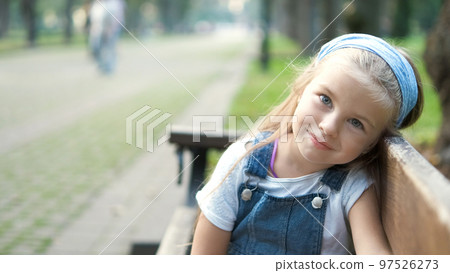 Small happy child girl sitting on a bench resting in summer park. Small happy child girl sitting on a bench resting in summer park. 97526273
