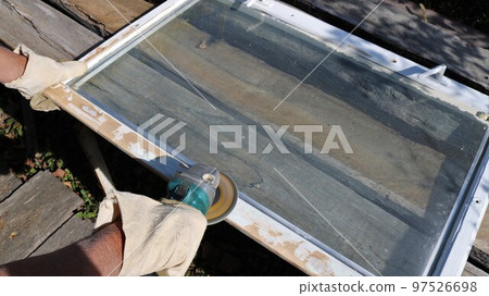 sanding a white-painted wooden frame, scraping off old paint and smoothing the surface of the wood with a grinder in the hands of a carpenter in canvas mittens in a street workshop 97526698